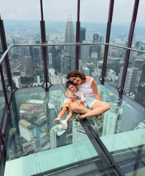 Mariangels y su hijo abrazados sobre el suelo de cristal de un mirador, con los rascacielos de Kuala Lumpur debajo